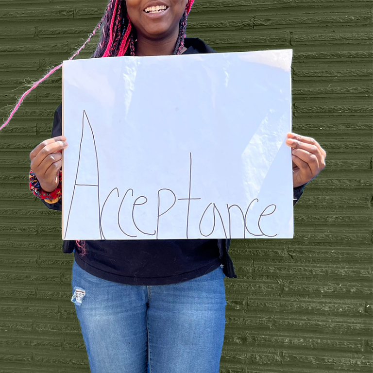 Woman at Grit Into Grace holds a sign that says acceptance as she stands in front of a green brick wall