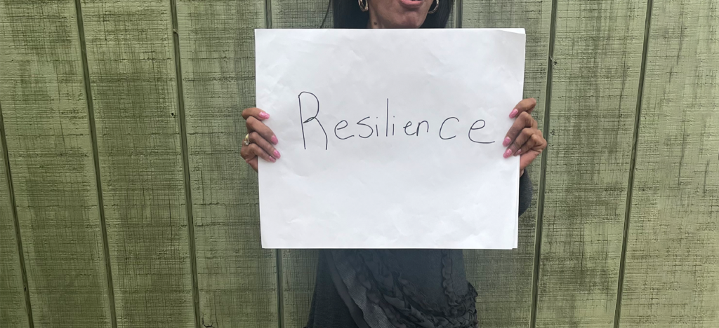 A woman from Grit Into Grace stands in front of a green wood wall holding a sign that says resilience