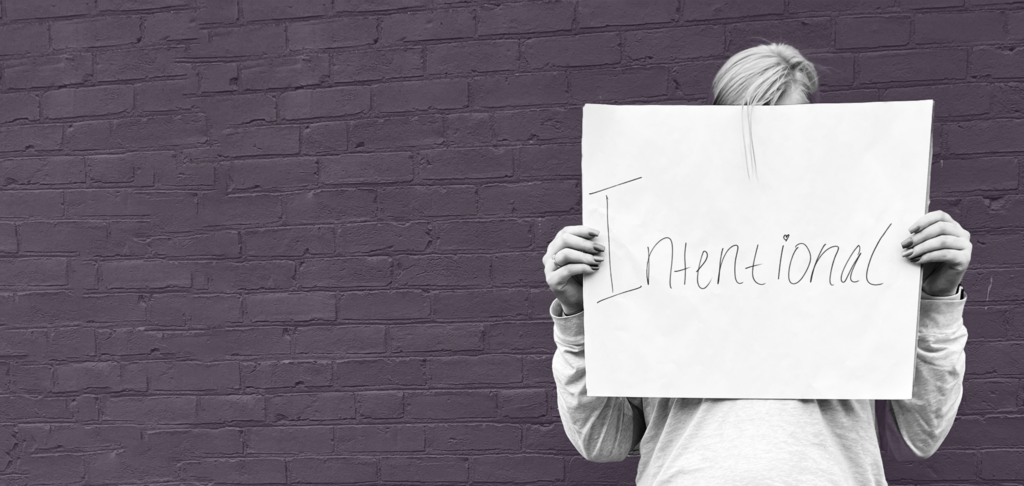 Woman from Grit Into Grace stands in front of a purple brick wall holding a sign that says intentional